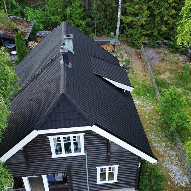 Luftfoto av mørkt hustak omgitt av trær og hage, med synlig veranda og tilbygg.